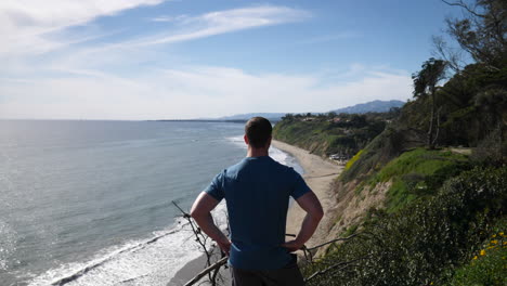 un hombre parado al borde de un acantilado oceánico en una ruta de senderismo natural con una vista épica de la playa en un día soleado en cámara lenta de santa bárbara, california