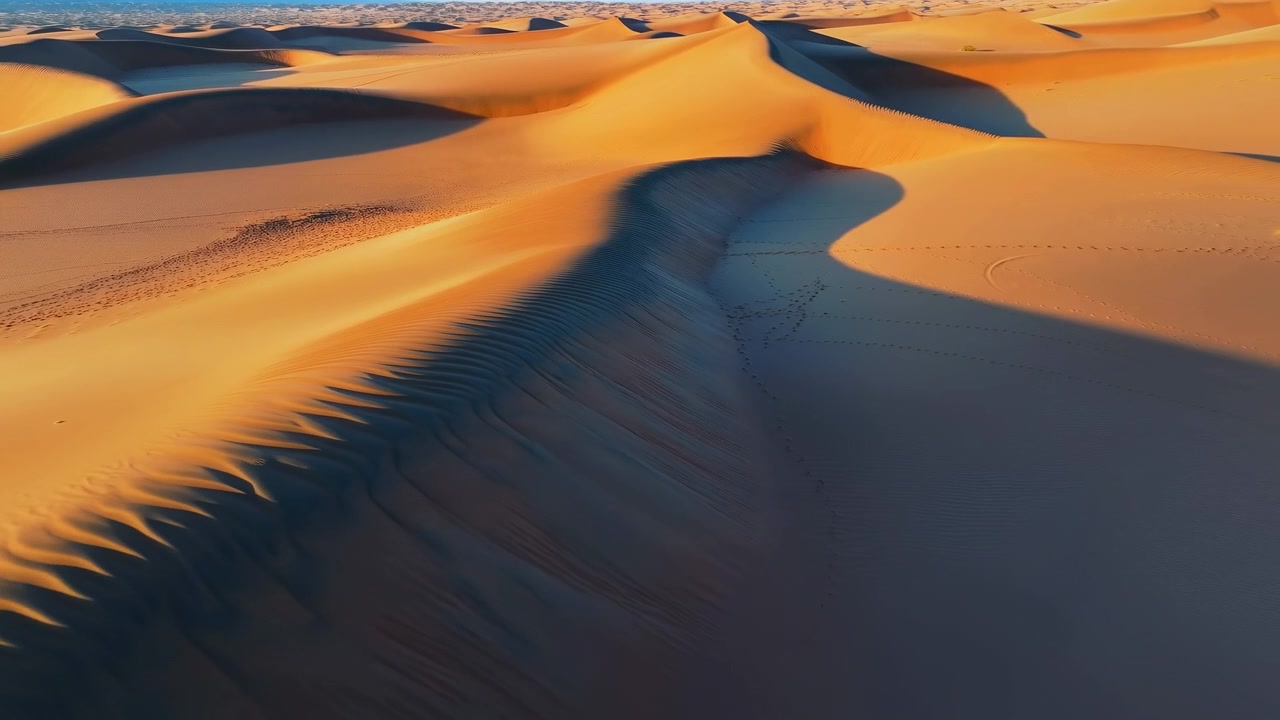 Vista aerea di dune desertiche ondulate modellate dalla calda luce solare e da profonde ombre blu, con una cresta principale che attraversa l'inquadratura e un vasto e tranquillo paesaggio sottostante