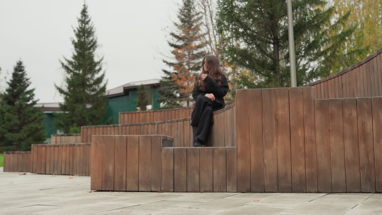 Ung kvinnelig student sitter på et amfiteater i tre, høstpark i bakgrunnen med furutrær, ryggsekk ved siden av seg, iført mørk frakk, leser i en notatbok med et ettertenksomt uttrykk, rolig overskyet lys, urbant
