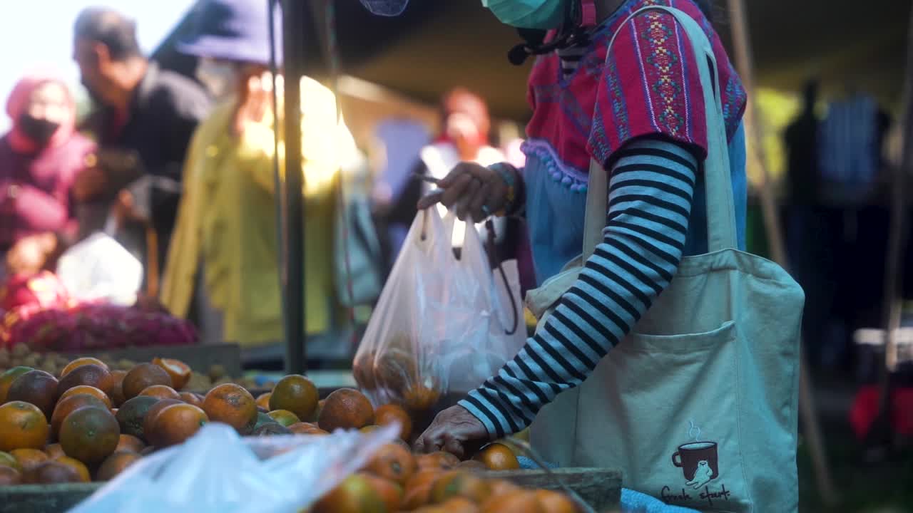 mujer asiática elige las naranjas más sabrosas con sus manos desnudas en el mercado tailandés local en pai en un día soleado mientras usa una máscara facial