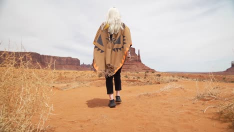 Young-woman-tourist-wrapped-in-brown-orange-blanket-with-traditional-pattern-with-blond-hair,-steps-along-path-at-Monument-Valley-landscape