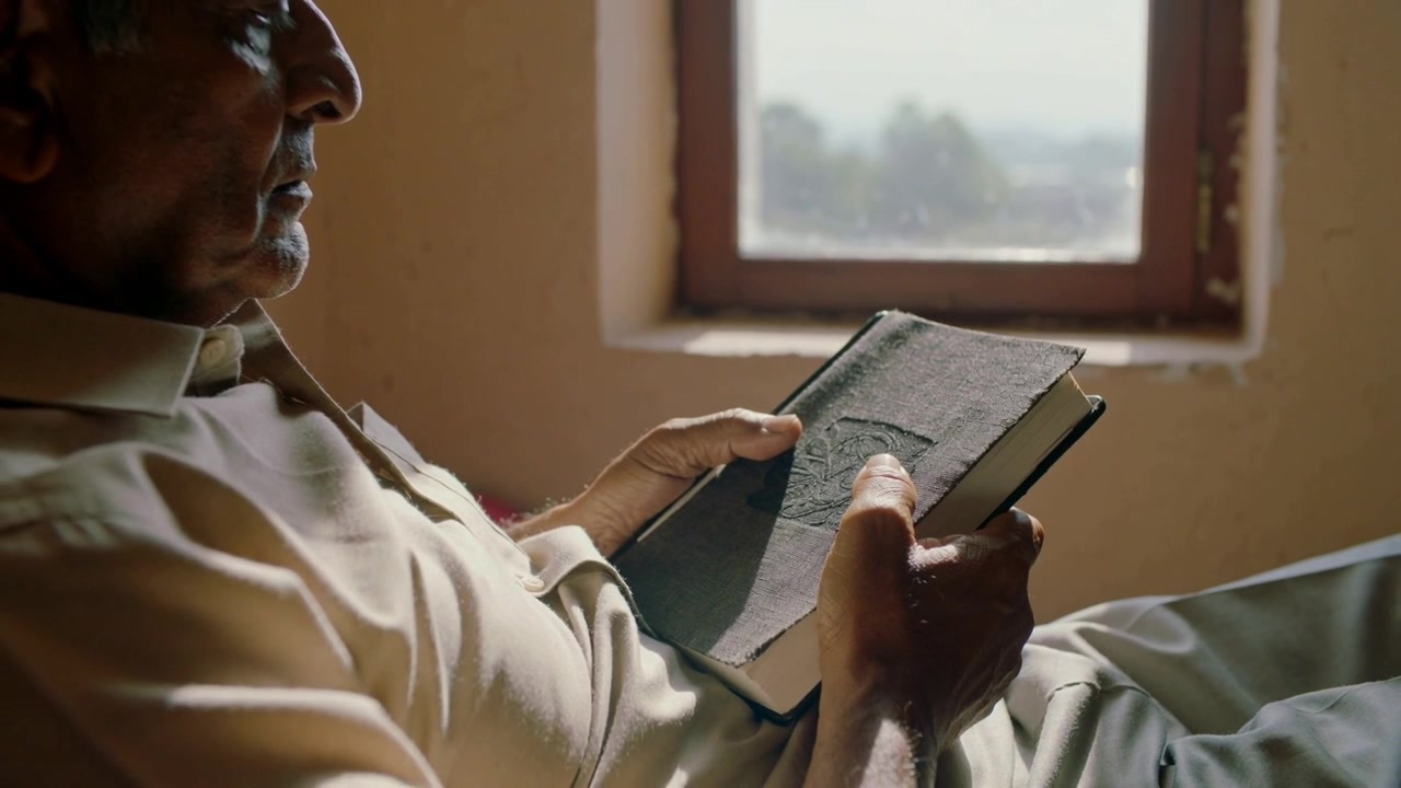 Camera pulling back showing senior man tracing embossed book cover at small room window, copy space