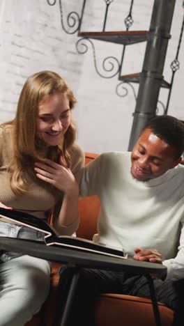 pretty couple of young white woman and positive african-american guy looks through old family photo album in living room at date slow motion