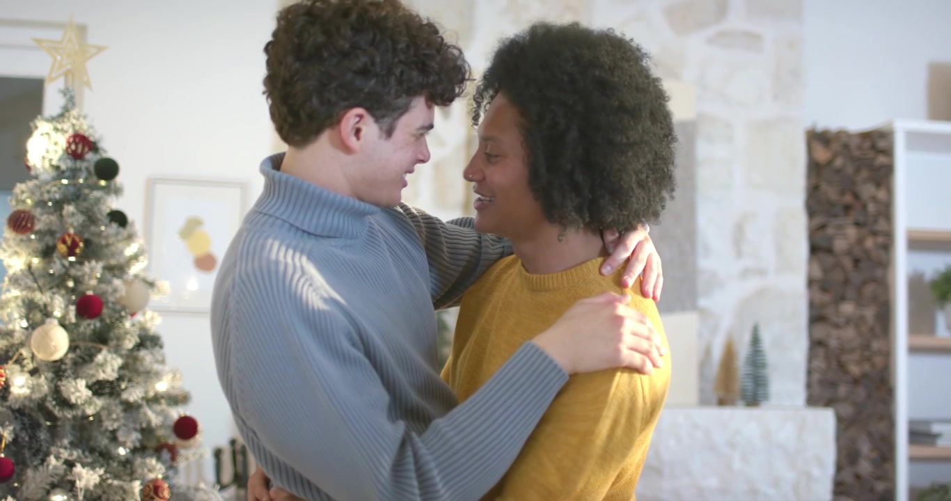African American men facing, moving closer and embracing by Xmas tree and fireplace during holiday