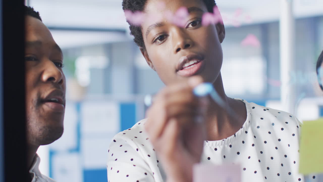African american business people brainstorming and writing on glass board at office, slow motion