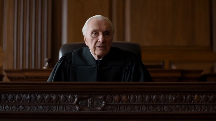 Anziano giudice bianco su una panca ornata, che pronuncia una sentenza solenne in un'aula di tribunale rivestita in rovere, con toga nera.