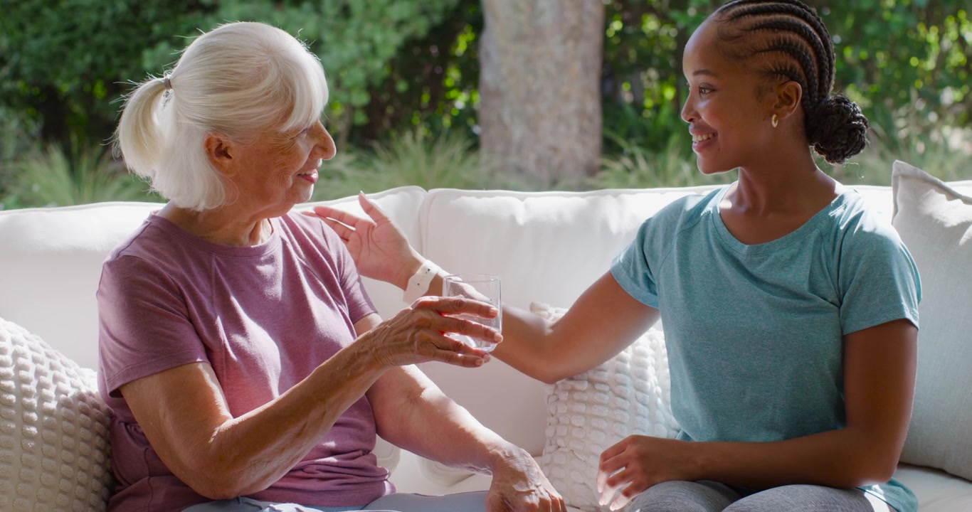 Aide-soignante afro-américaine tendant un verre d'eau à une femme âgée sur un canapé de patio, hydratant la personne âgée.