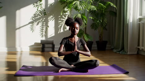 young black woman meditating in lotus pose on yoga mat in a sunny room