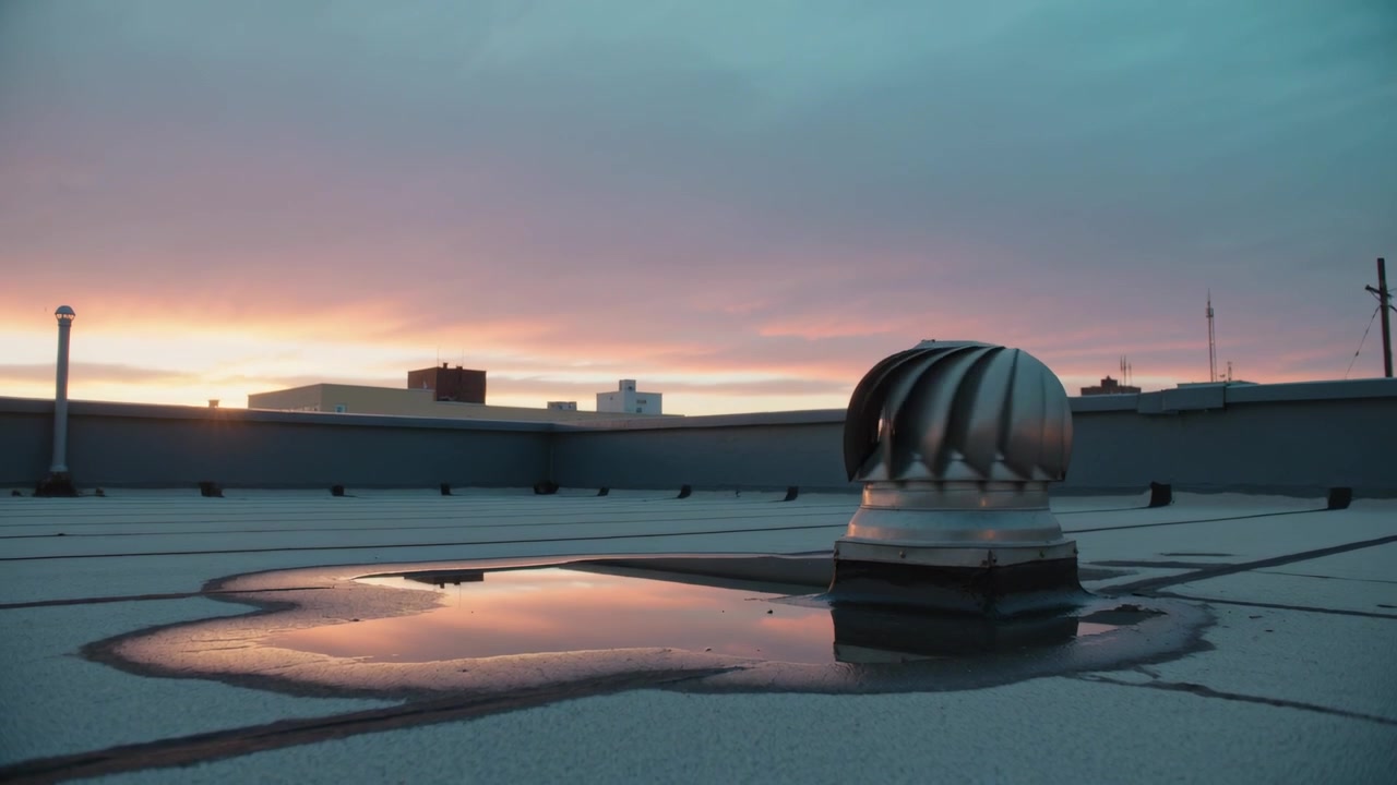 Turning metal turbine vent spinning in breeze on flat roof, puddle mirroring pink sky, copy space