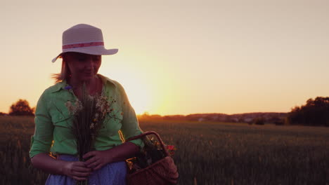 woman in a hat with a bouquet of wild flowers walking around the field at sunset 4k video