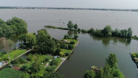 Toma-Aérea-En-Cámara-Lenta-De-La-Verde-Campiña-Holandesa-Con-Botes-Pequeños-Y-Lago,-Durante-El-Clima-Soleado-Y-Una-Pequeña-Ciudad-En-El-Fondo