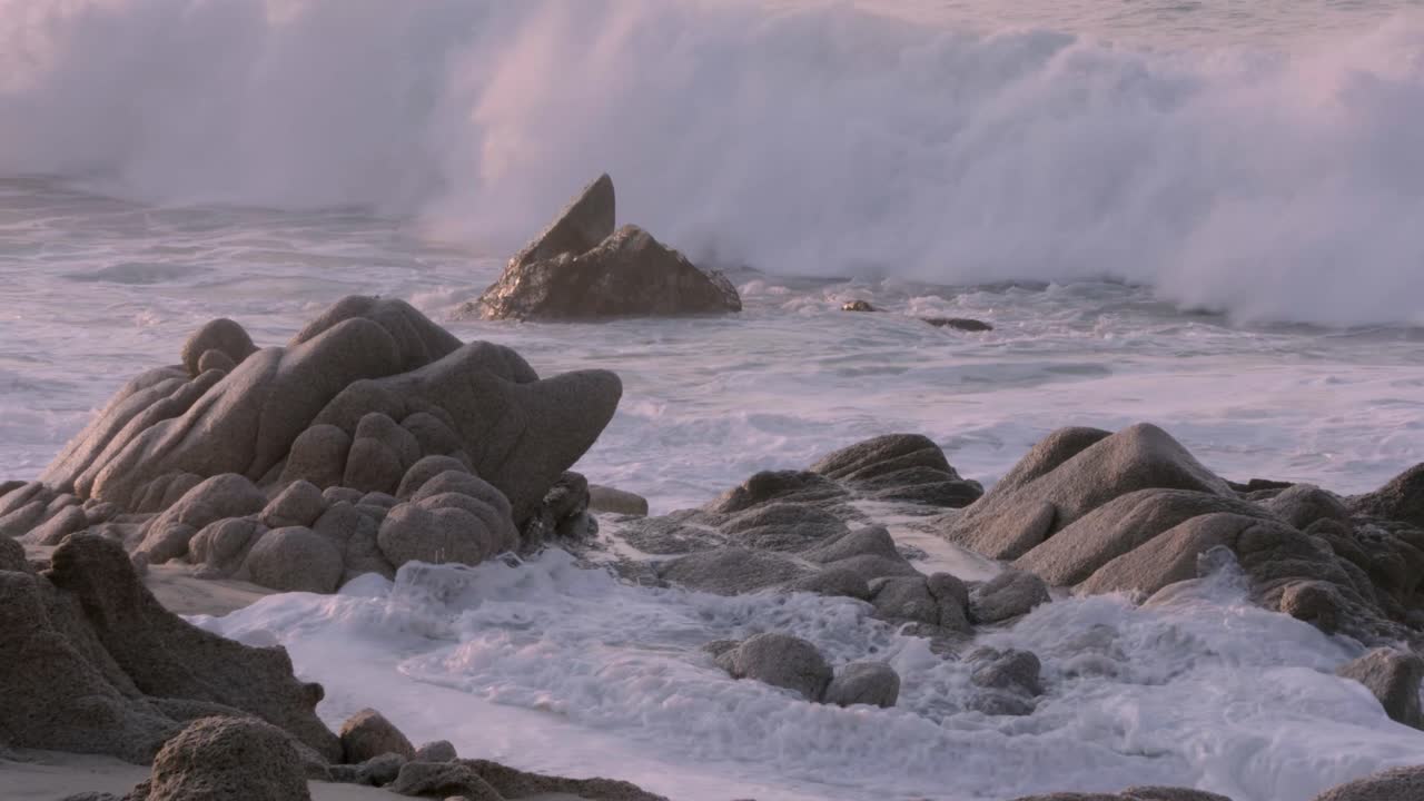 Wide shot of a sandy beach with waves crashing on large rocks, focused on the rugged coastal landscape under a calm, pastel sky. Slowmotion