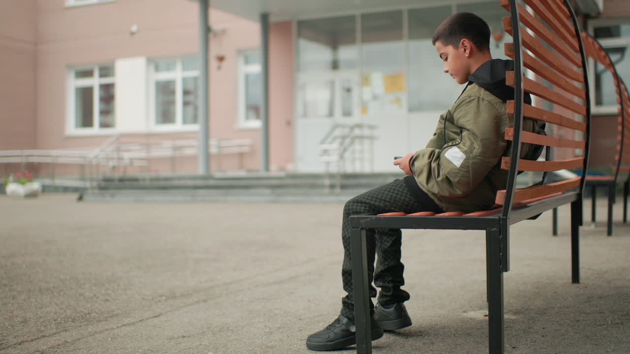 학교 건물 근처 나무 벤치에 앉아 휴대폰에 집중하고 있는 아이와, 차분한 분위기의 야외 풍경을 연출하는 문기둥 배경의 전구 불빛, 그리고 현대적인 건축물