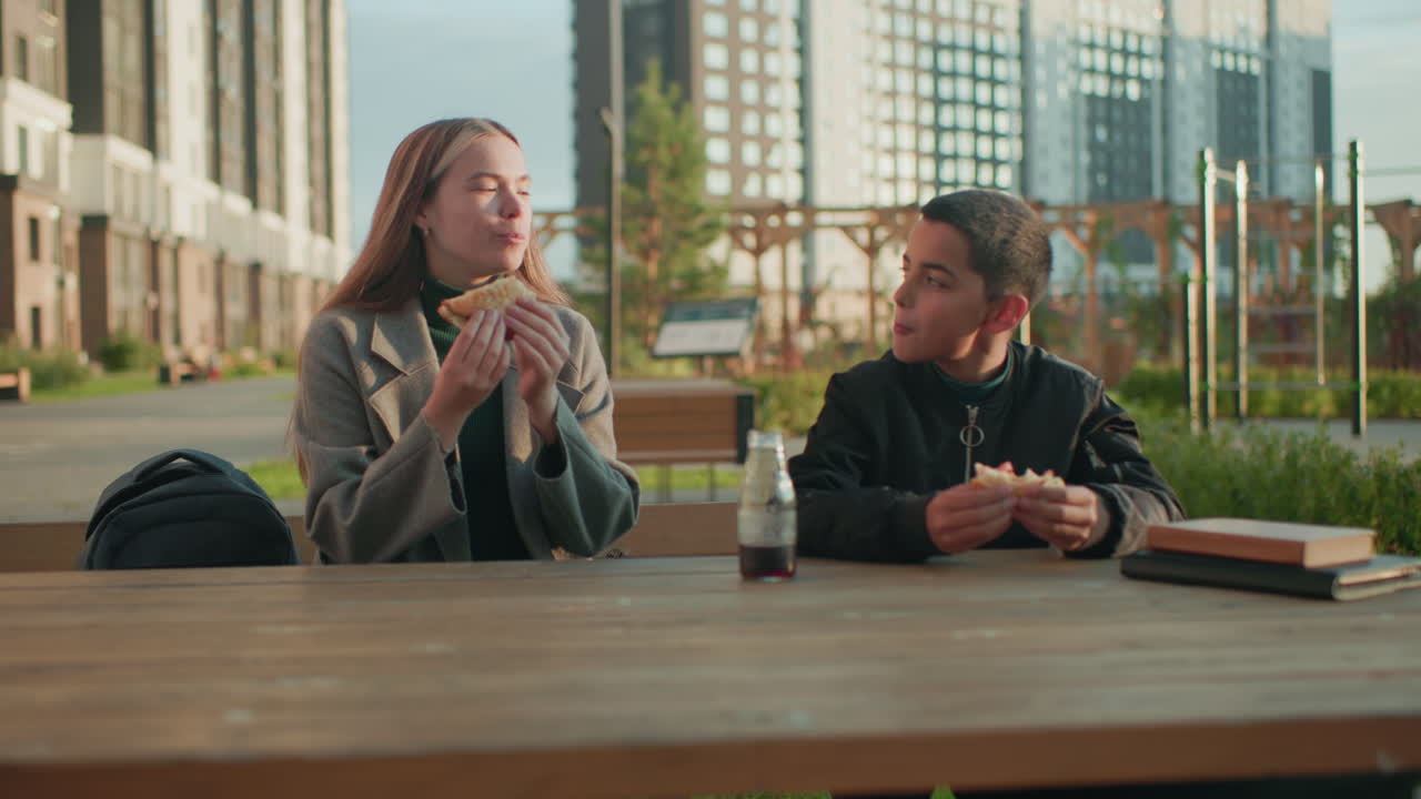Sister and brother sitting outdoors at wooden table in modern urban park making toast with pizza slices in hand, sharing happy snack time together with juice bottle placed on table in warm sunlight