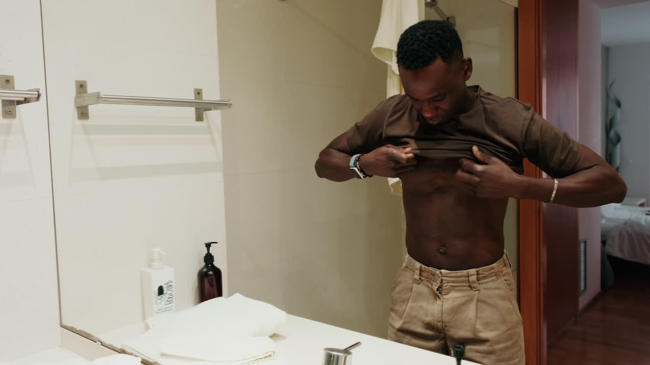 Young Black Man Putting on T-Shirt in Bathroom Mirror