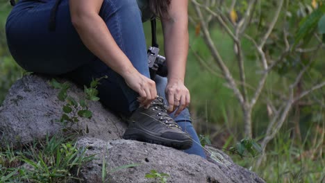 Female-explorer-with-her-backpack-and-camera-tying-her-shoelaces-while-preparing-for-a-hike-in-the-forest
