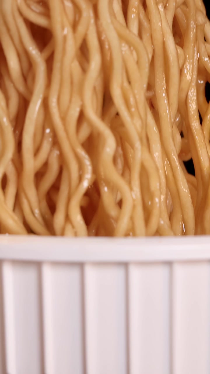 Primo piano al rallentatore di bacchette che sollevano noodles istantanei fumanti da una tazza bianca, con illuminazione drammatica.