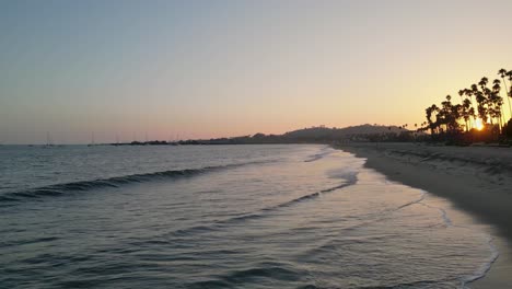 Drone-flying-up-Santa-Barbara,-CA-coastline-at-sunset-with-footage-of-ocean-waves-crashing-gently-against-the-beach-and-people-walking-on-the-shoreline