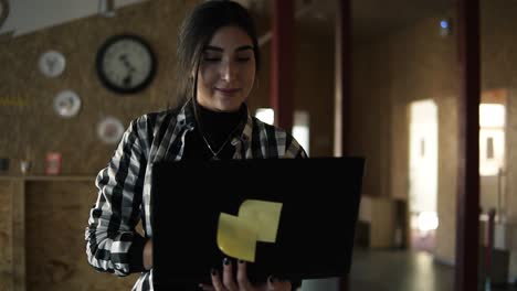 Close-Up-Beautiful-Young-Brunette-In-Plaid-Shirt-Is-Working-In-Modern-Workspace,-Walking-By-Room-With-Laptop