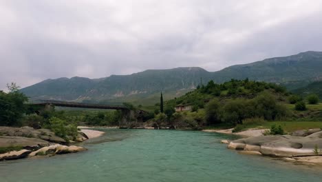 A-beautiful-river-flows-through-lush-greenery-in-Albania,-with-a-distant-mountain-range-and-an-old-bridge