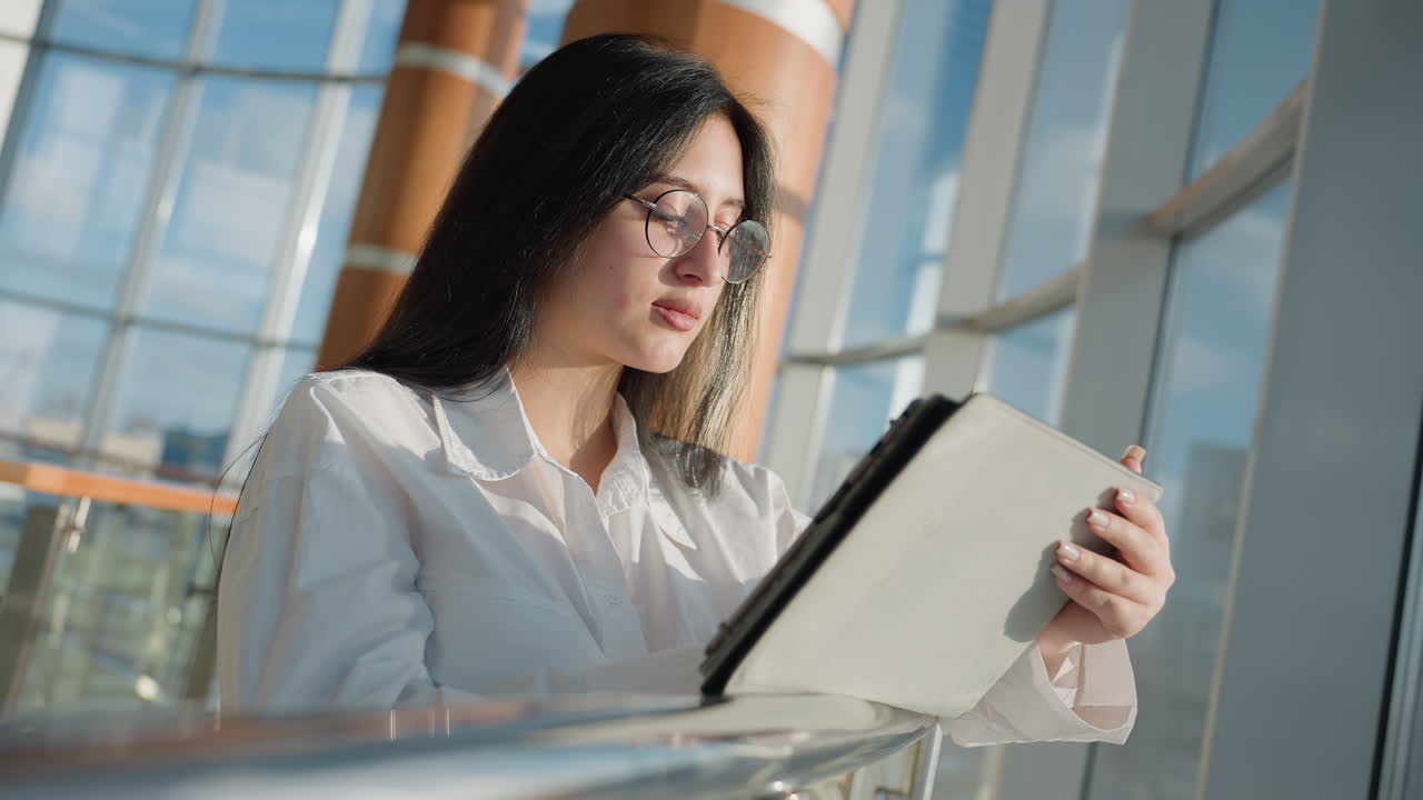 Elegant lady in white shirt holds tablet while sunlight gently reflects on her face, standing beside large glass window with modern cityscape view and natural light filling bright indoor space