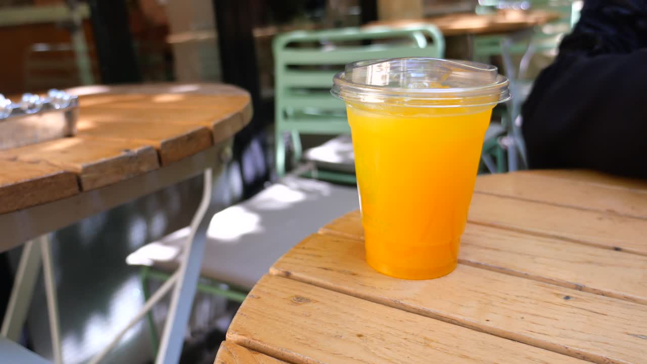 A hand reaching for a plastic cup of orange juice on a wooden table at an outdoor cafe