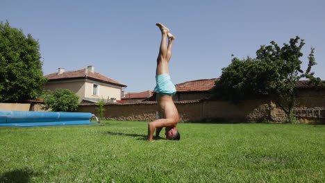 shirtless man in ashtanga headstand yoga pose on garden lawn