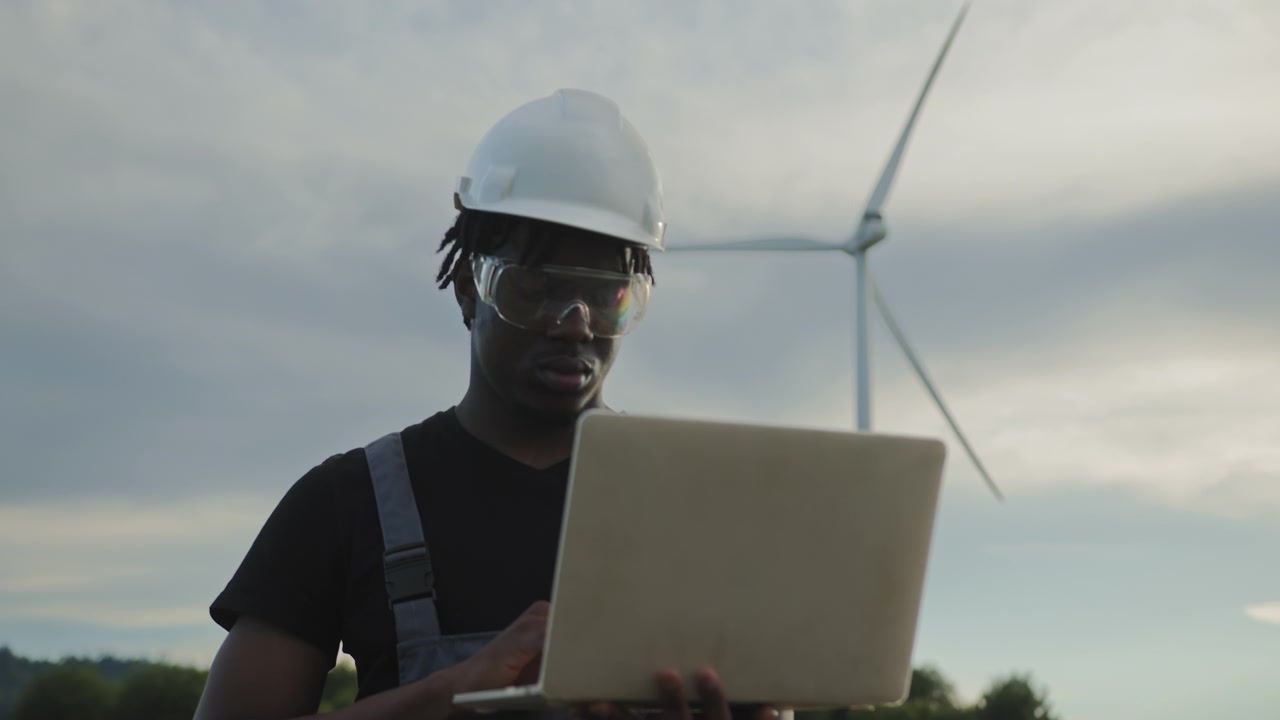 Man in hard hat and safety glasses uses laptop near wind turbine in field monitoring renewable