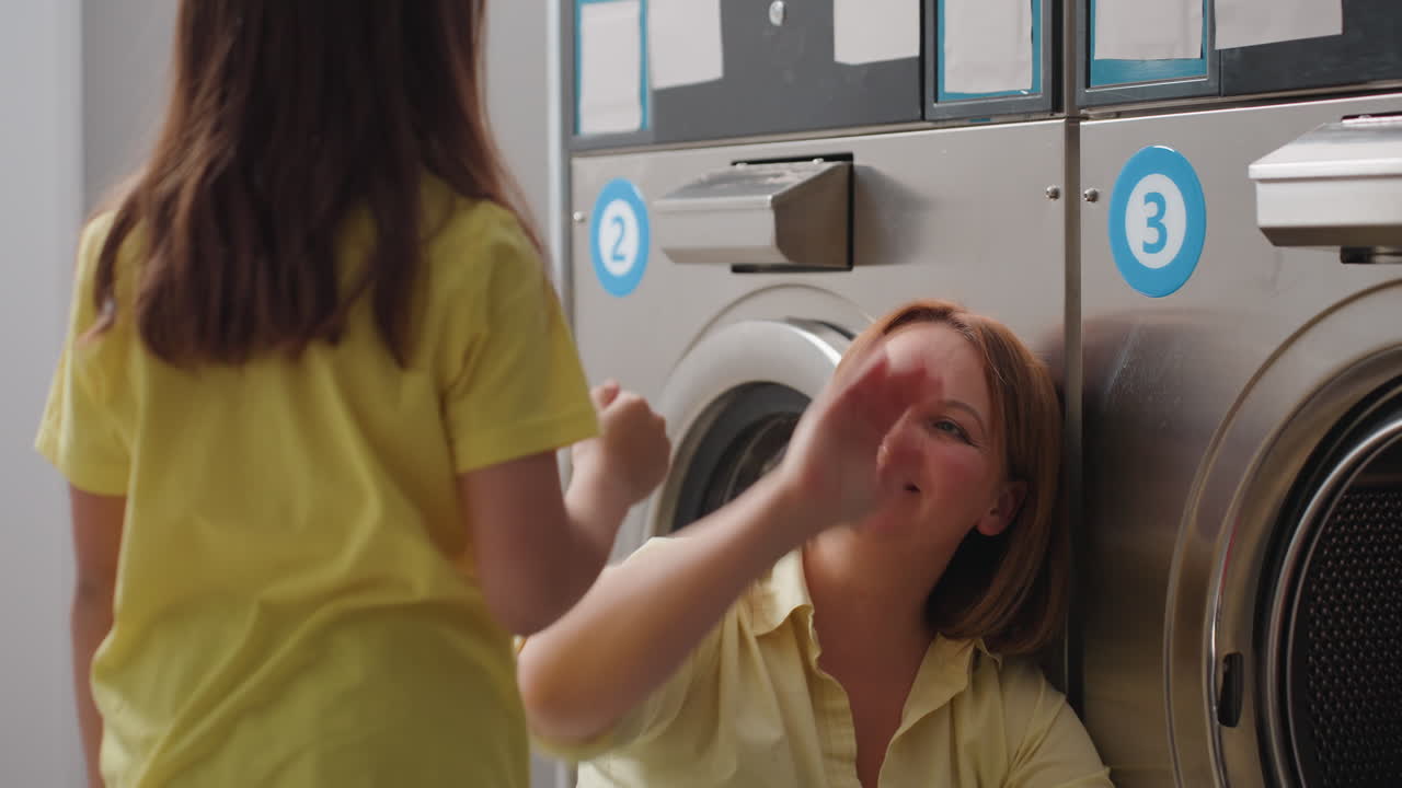 Cheerful mom seated by washer loads fabrics while smiling helper girl swings towel, meets palm for high five, bright laundromat shows teamwork, care, playful help during busy laundry task