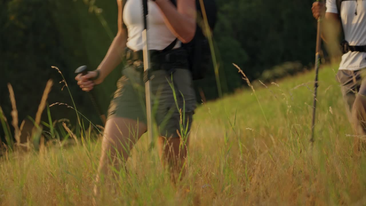 A couple hiking with trekking poles in a grassy field during sunrise, only their lower bodies visible