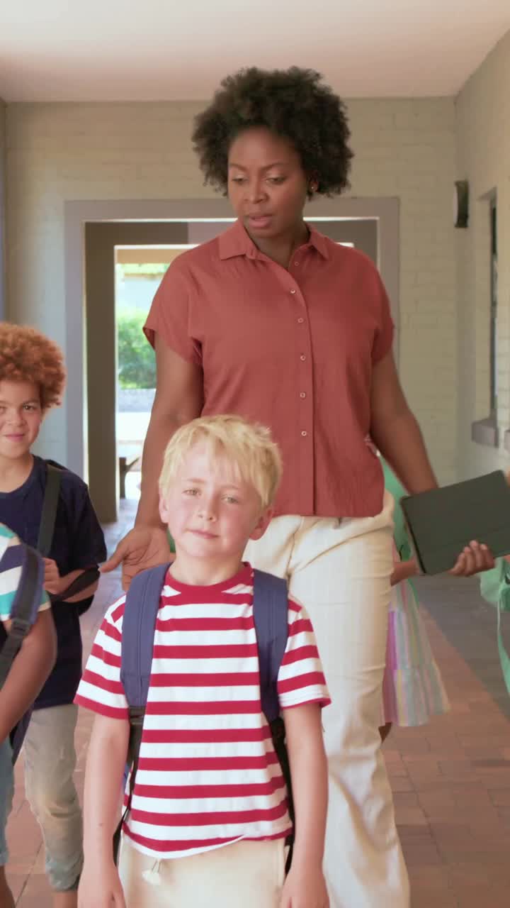 Vertical video: Entering teacher in rust shirt holding tablet guiding kids with backpacks at school