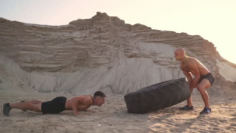 Two-athletes-train-in-active-mode-on-the-beach-doing-push-UPS-and-pushing-a-huge-wheel