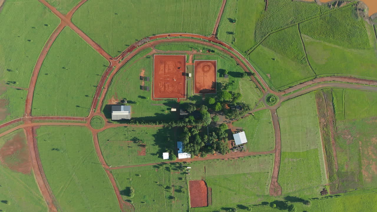 Amazing top-down aerial view over a rural horse riding area with green pastures and central red sand arenas.
