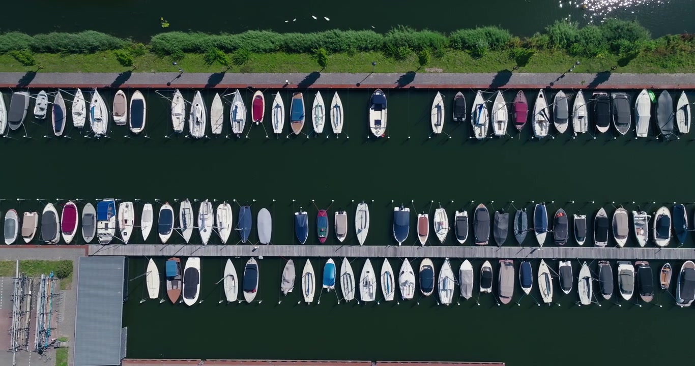 Droneoptagelse ovenfra, der viser små både fortøjet i pæne parallelle rækker, hvilket skaber slående symmetri ved siden af et parkeringsområde ved søen med krystalklart vand på en solskinsdag.