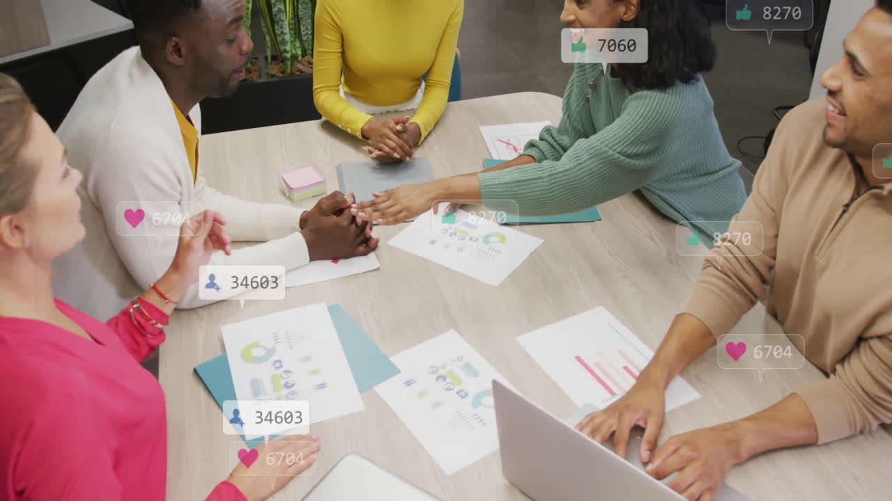 Collaborating at table, diverse team using animation with social media icons overlay