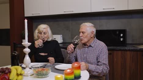 Portrait-of-senior-old-couple-toasting-wine-at-home-during-romantic-supper-in-kitchen-at-home