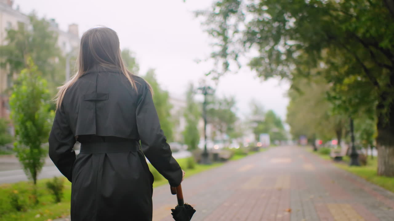 Woman in black trench coat holding closed umbrella walks along city street on overcast day, passing trees and parked cars, embodying autumn solitude, urban lifestyle, and moody seasonal atmosphere