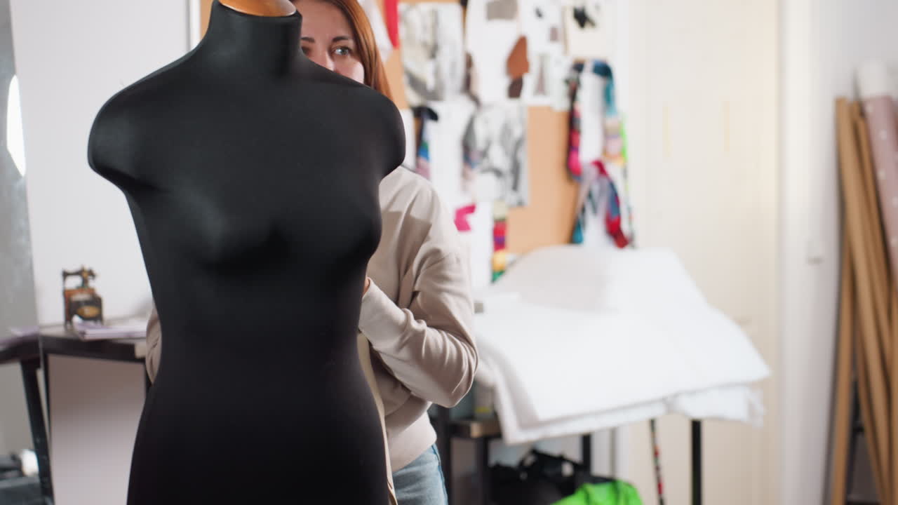 Creative designer removes blue velvet blazer from mannequin, steps back smiling with playful excitement inside fashion studio filled with fabric samples, inspiration sketches pinned on bulletin board
