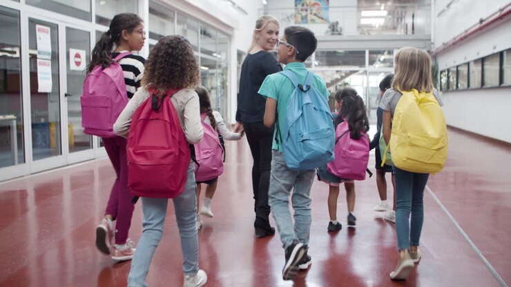 Vista trasera de niños multiétnicos caminando por el pasillo con una maestra.