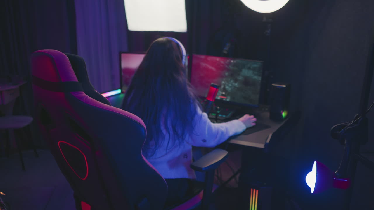 back view of woman with long hair wearing headphones sitting on gaming chair playing intense video game on dual screens in moody LED lit room with colorful lighting, mic, and pro studio gear setup