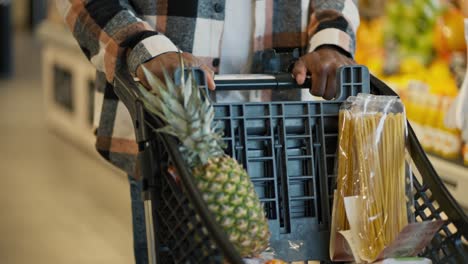 Close-up-shot-of-a-man-with-Black-skin-in-a-checkered-shirt-carrying-the-products-he-needs-into-a-cart-during-his-shopping-in-a-modern-grocery-store