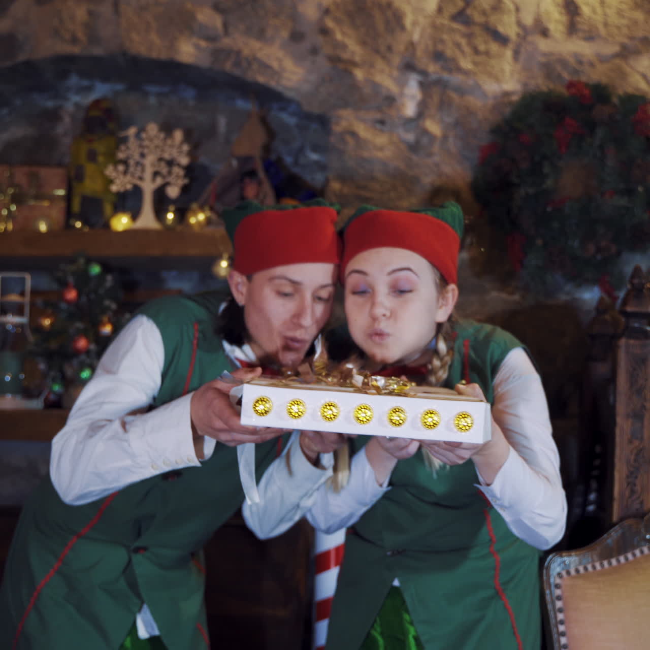 Joyful elves with magic book in decorated room at Christmas. Two elves in green costumes holding beautiful box and blowing out sparkles from top.