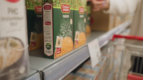 female customer uses fingertips to slide boxed cornflakes from shelf straight into wire cart, carefully aligning cereal packs among groceries while standing in brightly lit supermarket aisle