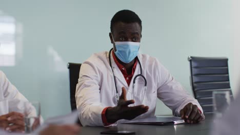 African-american-male-doctor-sitting-at-table-in-meeting-room-wearing-mask-speaking