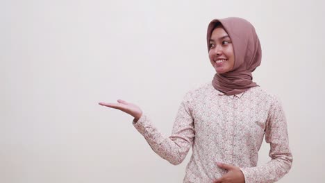 Happy-young-asian-muslim-woman-standing-while-presenting-something-beside-her-on-empty-space