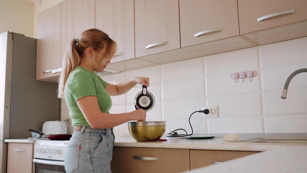 Woman in green shirt and jeans pours water into metal bowl while stirring ingredients during baking preparation on kitchen counter with utensils and appliances in background