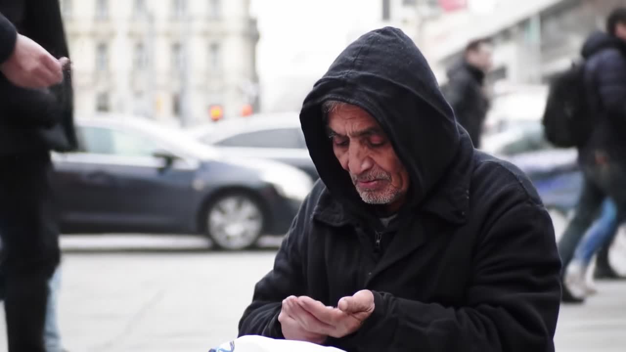 An older man wearing a hooded jacket sits on a busy urban sidewalk. He appears to be focused on something in his hands while a dog lies beside him. People pass by in the bustling backdrop.