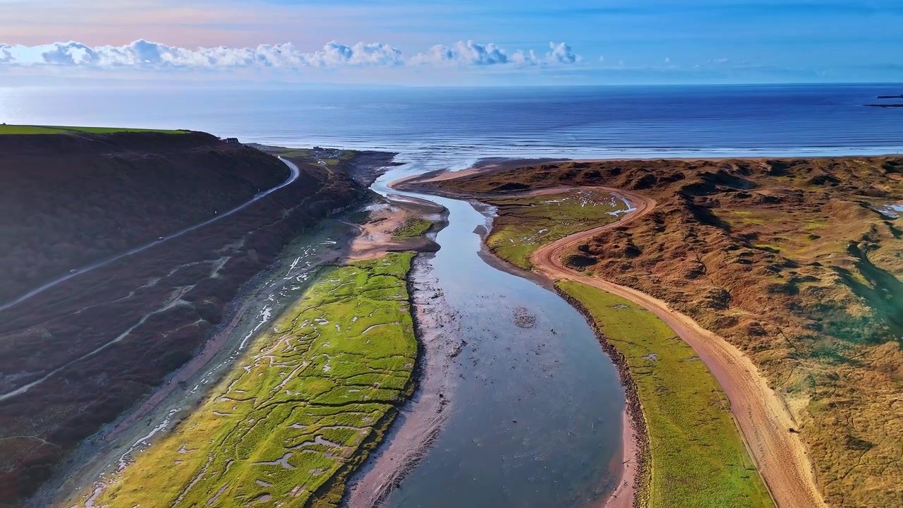 Veduta aerea della foce di un estuario fluviale che si apre verso il mare al crepuscolo. Un estuario fluviale si snoda verso il mare attraverso dune di sabbia e palude salmastra, mentre un cielo dai colori pastello delinea l'orizzonte al crepuscolo.