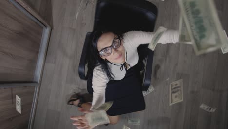 top view cheerful, smiling and a little crazy young brunette in white blouse and black skirt spinning on office chair. happy  manager or secretary. dollars are falling on her  from above