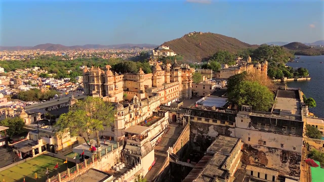Una ripresa statica grandangolare dall'alto che mostra il maestoso complesso del City Palace sulle rive del lago Pichola, con il paesaggio urbano e le colline Aravalli sullo sfondo, Udaipur, Rajasthan, India.
